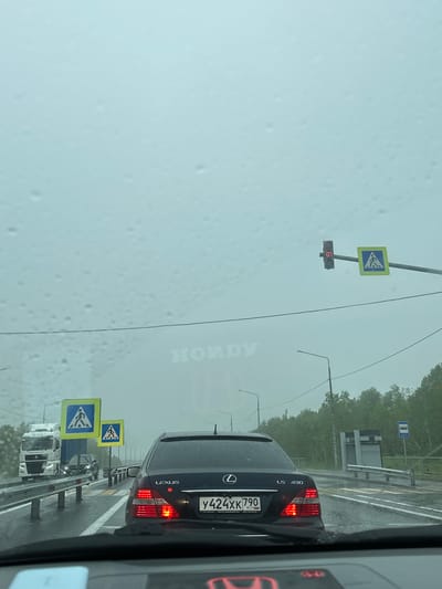 The image depicts a rainy day on a multi-lane road in Pionersky, Russia. The perspective is from inside a vehicle, looking forward. A black Lexus LS 430 is in the foreground, with its rear lights illuminated. The road surface is wet, indicating recent rainfall, and water droplets are visible on the windshield.
Several traffic signs are present. Two pedestrian crossing signs, one larger than the other, are visible on the right side of the road. Further ahead, a traffic light with a red display showing "07" is illuminated.
In the distance, a white truck is on the left lane. The surrounding environment features dense green trees and a grey, overcast sky. The overall atmosphere suggests a traffic situation during inclement weather.