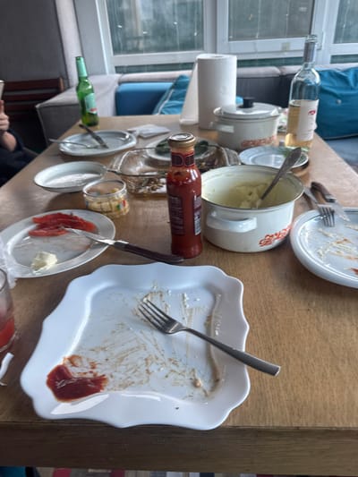 The scene depicts a dining table in Ptichnoye, Russia, covered with the remnants of a meal. Several empty plates, some with food residue, are visible, alongside used silverware, including forks, knives, and spoons. A white enamel pot containing mashed potatoes sits near a partially full bottle of ketchup. Other containers hold unidentified food items. Bottles of wine and a paper towel dispenser are also present. A window is visible in the background, and a sofa with cushions. The focus is on the disordered state of the table after a meal, with no people clearly visible, only a partial view of someone's clothing in the lower left.