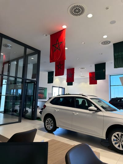 The image shows the interior of a Škoda dealership in Ouahat Sidi Brahim, Morocco, possibly during a promotional event. Red and green Moroccan flags, some emblazoned with the Škoda logo, hang from the ceiling. A white Škoda vehicle is partially visible, occupying the right side of the frame. In the lower portion of the image, a modern glass-enclosed area is visible, showcasing advertising displays related to Škoda. A black chair and a computer monitor are present in the foreground. The ceiling features several ventilation grilles and recessed lighting.