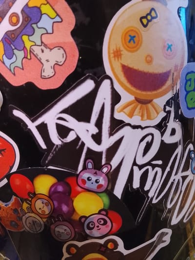 The image depicts a collection of stickers adhered to a black surface. The stickers include a stylized brain, a ragdoll-like face with buttons for eyes and a bow, a plate of fruits and animal-faced candies, and a large signature written in white. The location is Высокиничи, Russia.