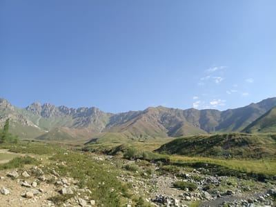 The image depicts a panoramic view of a mountainous landscape under a clear blue sky with scattered wispy clouds. The foreground is a rocky, uneven terrain covered in dry grasses and scattered stones. A narrow, shallow stream or dry creek bed runs diagonally through the lower right portion of the image, also filled with stones. In the midground, rolling green hills and sparse vegetation dominate, with some areas appearing drier and more sparsely vegetated. Further in the background, rugged, rocky mountains rise sharply, their slopes showing variations in color from green to brown and grey, with some patches of snow or scree visible on the higher peaks. The overall impression is one of a remote, natural environment in a mountainous region. The scene does not show any people, animals, or man-made structures, suggesting a wild or unpopulated area.