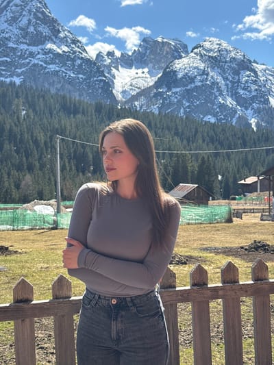 A woman is sitting on a wooden fence with snow-capped mountains in the background in Sappada, Italy. The woman has long brown hair and is wearing a gray long-sleeved shirt and jeans. She is looking to the left, away from the camera. The background shows a lush green forest and a bright blue sky with some clouds. There are buildings in the distance. The woman is posed near a wooden fence in the foreground.