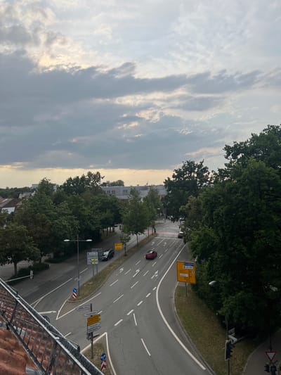 The image depicts a street intersection in Straubing, Germany, on an overcast day. The view is from an elevated perspective, looking down on a road with multiple lanes and distinct traffic markings. A red car is visible in the left lane, and a dark colored vehicle is in the right lane. Several road signs are present, including directional signs pointing to "Polizeipräsidium," "Regensburg," "Geiselhöring," "Tiergarten," and "Westpark." There are also signs indicating parking areas and a speed limit of 30 km/h. Mature trees line the right side of the road, and buildings are visible in the background. A metal railing and terracotta roof tiles are in the foreground, suggesting the vantage point is from a balcony or rooftop. The sky is filled with scattered clouds, with some brighter areas indicating the sun attempting to break through.