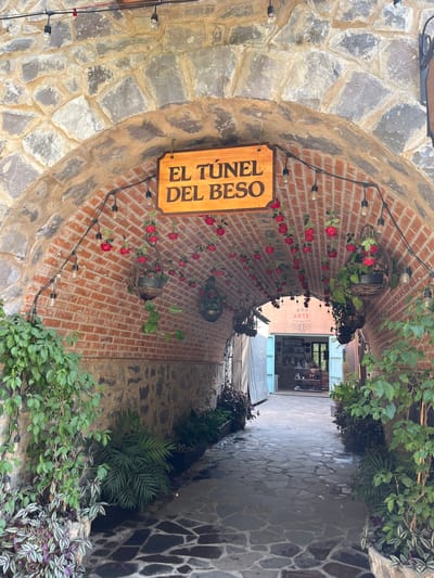 The image depicts a stone tunnel entrance, presumably in Domingo Arenas, Mexico, leading into a narrow passageway. A wooden sign reading "El Túnel Del Beso" is prominently displayed on the left side of the entrance. The tunnel's archway is constructed of stone and brick, with decorative red roses and hanging planters adorning the brick walls. String lights are also present along the arch. The passageway beyond the tunnel entrance is paved with irregular stone slabs, and plants grow along the walls. In the distance, a storefront with the word "ARTE" partially visible is seen at the end of the passageway. There are no people visible in the image.