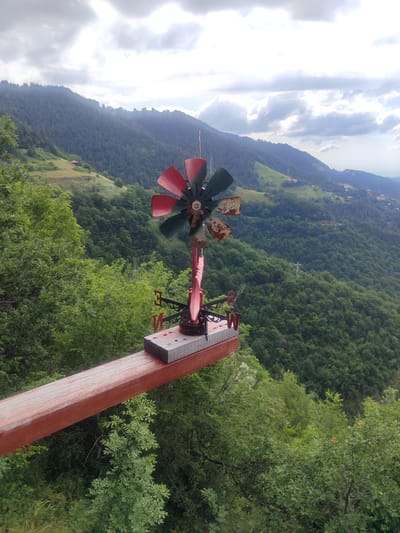 This is a medium shot, eye-level, outdoor photograph of a weather vane on a wooden plank overlooking a lush green mountainous landscape. The weather vane is in the foreground, positioned on the right side of the frame. It features a red and green propeller with a rusty center and a red pointer with the letters "E" and "W" on either side. The wooden plank it rests on is red and has some wear and tear.

In the background, rolling hills covered in dense green trees stretch into the distance. The sky is overcast with dramatic clouds, casting a diffused light on the scene. The landscape appears to be in or near the city of Pechinska, Bulgaria, given the context. No people are visible in the image. The overall impression is one of natural beauty and rustic charm.
