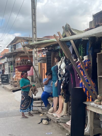 The image shows a street scene in Isheri, Nigeria. A woman is walking towards the right, carrying a large basket filled with food items on her head, and she is wearing a green shirt, a patterned skirt, and sandals. A second person, wearing a blue shirt and pants, is seen from the back and is facing towards the left. Market stalls line the street, showcasing various goods, including clothing and mannequins. The stalls are constructed with wood and covered with tarps, and some items hanging from the stalls. A sign indicating "OFAJA RICE SPOT" is visible. The street appears to be paved, and the sky is overcast. A concrete structure runs across the top left of the image.