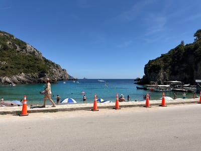 The image depicts a sunlit coastal scene in Palaiokastritsa, Greece, characterized by a blue sky, clear turquoise water, and verdant cliffs.

In the foreground, a light grey asphalt road occupies the bottom third of the frame. Six bright orange traffic cones, each resting on a black rubber base, are evenly spaced along the road's edge, positioned on a narrow, light-colored concrete strip. This strip abuts a low, light-colored stone wall, approximately 0.5 to 0.7 meters high, which separates the road from the beach area.

On the beach and in the water's edge, a woman wearing a patterned dress and a wide-brimmed hat walks from left to right along the stone wall, carrying sandals in her left hand. Two beach umbrellas are visible: one with blue and white stripes to the left, and another with blue and yellow stripes further right. Approximately 10-15 individuals are present in the shallow, clear water, swimming, wading, or standing. A pink inflatable tube is visible near one individual in the water. A yellow floating rope with buoys defines a swimming area further out.

The midground features the calm, blue-green water of a cove. Several white motorboats are anchored or moored within the cove. On the far right, a small dock extends into the water, accommodating multiple larger tour boats, some with white canopies, and approximately 5-10 people are visible on or near this dock.

The background is dominated by steep, rocky cliffs covered in dense green vegetation, framing the cove on both the left and right sides. These cliffs open to the wider, deeper blue sea beyond. A small, dark island or rock formation is discernible on the distant horizon in the center. The sky above is a clear, bright blue with minimal, wispy clouds in the upper right. A faint white contrail from an aircraft is visible in the upper left quadrant of the sky.