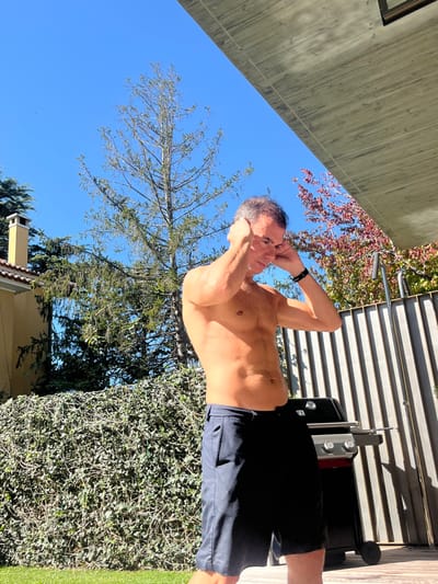 A shirtless adult male with dark hair and a tanned, athletic physique stands outdoors in a residential garden located in Lordelo do Ouro e Massarelos, Portugal. He is wearing dark-colored shorts and a black smartwatch on his left wrist. His hands are raised to the sides of his head, and his gaze is directed downwards and slightly to his right. The man is positioned on a patch of green grass. To his immediate left is a dense green hedge. To his right, behind him, is a dark-colored gas barbecue grill with silver accents, situated in front of a grey, vertical slatted fence. The background features several trees: a tall, sparse conifer, denser green foliage to the left, and a tree with reddish leaves to the right. A light yellow building with a chimney is partially visible on the far left. A clear blue sky dominates the upper portion of the image, with a concrete architectural overhang visible in the upper right corner. Strong sunlight illuminates the scene, creating distinct highlights and shadows.