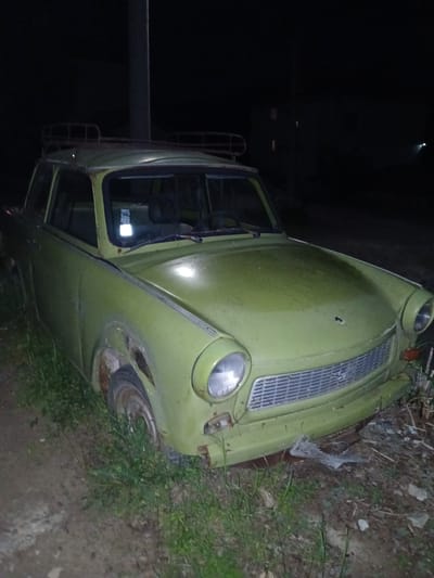 A partially obscured, vintage, olive-green car is located in Pripek, Bulgaria. The car appears to be a model from the mid-20th century. The vehicle is parked on a dirt and grassy verge on the side of the road. The car's exterior shows signs of age and wear, including rust. The photograph was taken in low lighting conditions, possibly at night, with a dark background. A metal pole is visible next to the driver's side door. The front of the car is facing the right side of the image frame.