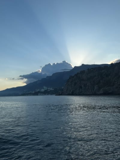 The image depicts a seascape with a rocky coastline. The sky is bright blue with visible sun rays emanating from behind a mountain or large rock formation. A body of water, likely the sea, occupies a significant portion of the right side of the image. The coastline is rugged, with dark, textured rock formations. There is evidence of habitation along the coastline, possibly buildings. The scene is located in or near Гаспринский поселковый совет, Ukraine, based on the prompt.