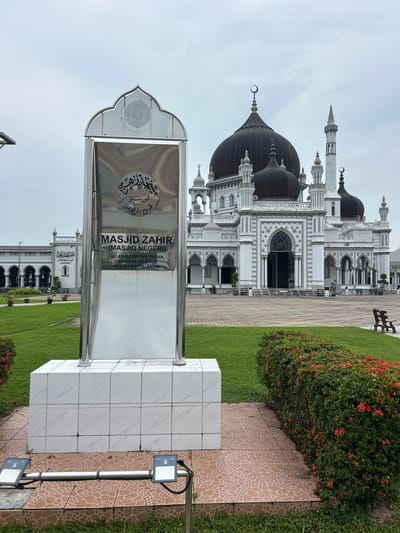 In Alor Setar, Malaysia, the Masjid Zahir, also known as Masjid Negeri, is depicted. A reflective metal monument stands in the foreground, bearing the mosque's name and address: Jalan Kampung Perak, 05100 Alor Star, Kedah Darul Aman. Behind the monument, the mosque presents an impressive facade with multiple dark domes and white minarets against a cloudy sky. The mosque's architecture is detailed, featuring arches, decorative elements, and a grand entrance. The surrounding area is landscaped with green grass, a paved walkway, and a dense hedge of flowering bushes. A wooden bench is visible in the middle ground. Two ground lights are positioned near the base of the monument. The scene is serene and appears to be captured on an overcast day.