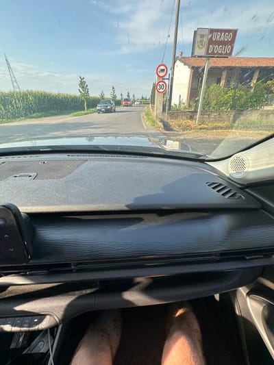 The image, taken from inside a car in Rudiano, Italy, shows a road with cornfields on the left and a building with a sign that reads "URAGO D'OGLIO" on the right. There are traffic signs indicating a speed limit of 30 and a restriction for trucks. Several cars are visible on the road. The car's dashboard is in the foreground. The sky is clear and blue with some clouds.