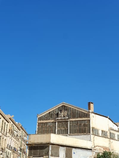 The image shows a clear blue sky above a building in Oran, Algeria, located at coordinates 35.704 latitude, -0.652 longitude. The building, constructed from tan concrete blocks, has a large glass window on the top level, consisting of multiple panes of glass, with a metal frame surrounding them. The top of the structure is covered in a metal roof. The building appears to be older, with some of the glass panes broken or missing. A smaller building appears below the main building, made of the same tan concrete blocks. This building has multiple windows and balconies that appear to be covered by awnings or curtains. There are no people visible in the image.