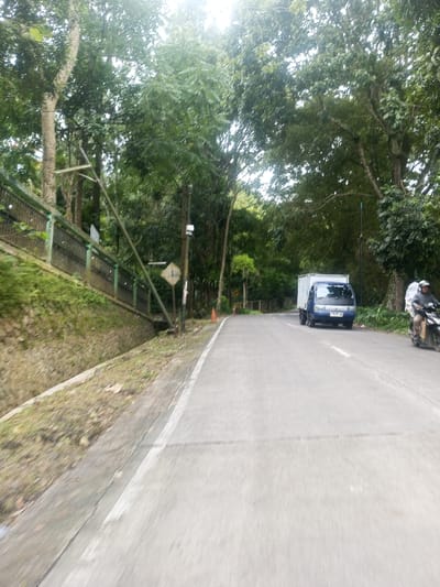 The image displays a paved road in Sarwahi Mal., India, lined by dense green trees and vegetation, under an overcast or diffused daylight sky. The road's surface is light gray, with a solid white line marking its right edge.
On the left side of the road, an earthen embankment rises, topped by a dark green metal fence. A concrete drainage ditch runs along the base of this embankment. A utility pole stands near the ditch, next to a significantly tilted light pole extending to the left. A yellow, diamond-shaped road sign with a black symbol is affixed to the upright utility pole, below a small white rectangular device. An orange traffic cone is visible on the paved shoulder further down the road on the left.
On the right side of the road, lush foliage and trees form a continuous border. A blue and white box truck, possibly a light-duty commercial vehicle, is observed traveling towards the viewer in the right lane. Its license plate is partially visible, displaying "L 9070 WA". To the right and slightly behind the truck, a person on a black scooter-style motorcycle is also traveling towards the viewer. The motorcyclist is wearing a light-colored helmet and a light-colored outer garment.