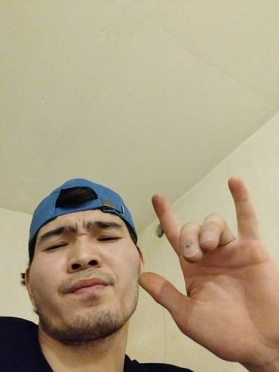 A young man wearing a blue baseball cap and a black t-shirt is shown in a close-up, low-angle selfie. He has a light beard and mustache and is making a "rock on" hand gesture with his right hand. His eyes are partially closed, and his facial expression appears to be one of amusement or playfulness. The background is a lightly textured, cream-colored wall and ceiling, indicating an indoor setting. There is no indication of the city of Tuzdybastau, Kazakhstan in the image.