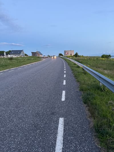 An asphalt road with white dashed lane markings curves towards the left, bordered by a metal guardrail on the right. Green grass and yellow wildflowers grow along the roadside. In the background, several buildings, including a modern house with a dark roof and a smaller utility building, are visible. Further in the distance, a round, stone ruin resembling a tower stands on a grassy hill. Two cars are on the road, one with its headlights on. The sky is a pale blue with scattered clouds, suggesting either early morning or late evening. The scene appears to be a rural or coastal area, possibly near Paldiski, Estonia, given the presence of coastal landscape elements and historical ruins.
