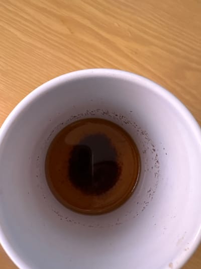 An overhead shot shows the inside of a white mug on a wooden table. The mug contains a small amount of dark brown liquid, likely coffee, with a ring of dark particles around the rim and a darker concentration of sediment at the bottom. The image does not provide any information about the city of Rodniki, Russia.