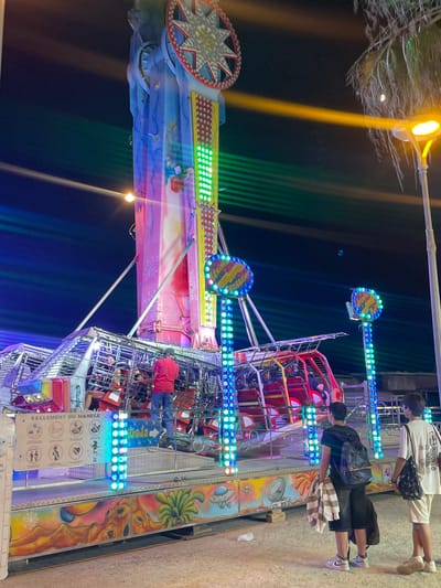 This media file captures a vibrant scene at a nighttime amusement park in Canet De Mar, Spain. The central focus is a brightly lit amusement ride, likely a thrill ride, with a towering structure adorned with colorful lights. The ride features multiple gondolas or carriages, some of which are occupied by people, their faces partially obscured by the lighting and angle of the photograph. The scene is illuminated by a mix of artificial lights from the ride itself, a yellow street lamp casting a strong beam, and green lights creating streaks in the dark background.
In the foreground and background, several individuals are visible. Some are standing near the ride, possibly waiting to board or having just disembarked, while others are seated within the ride's structure. The people are dressed in casual attire. One person in the background is wearing a white t-shirt and beige shorts, carrying a black bag. Another person, closer to the ride, wears a black t-shirt and black shorts, also with a bag.
The overall mood appears lively and festive, characteristic of a fairground at night. The numerous lights and the presence of the amusement ride suggest an event or attraction is in full swing. The ground appears to be paved, and some foliage is visible in the upper left corner, hinting at an outdoor setting. The time of day is clearly night, indicated by the dark sky and the prominence of artificial illumination.
Visible text on a sign attached to the ride is in French, reading "REGLEMENT DU MANEGE" which translates to "RIDE REGULATIONS." Below this, there are several pictograms illustrating rules or warnings, such as "Ne pas se pencher dehors" (Do not lean out) and "Tenir les enfants serres" (Hold children tight).