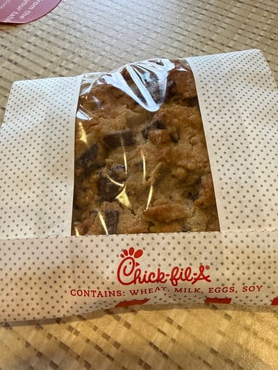 The image shows a packaged chocolate chip cookie from Chick-fil-A. The packaging is primarily white with gray polka dots, and a transparent section revealing the cookie inside. The Chick-fil-A logo is printed in red on the packaging, along with an ingredients list: "CONTAINS: WHEAT, MILK, EGGS, SOY." The cookie appears to be enclosed in clear plastic within the packaging. The package sits on a surface with a grid pattern. A small portion of a pink circular object is visible in the lower left corner, marked with the words "from the your tabl Code."