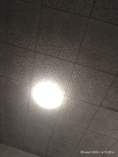 The image displays an upward-angled view of an indoor ceiling, purportedly located in Balgarsko Slivovo, Bulgaria, recorded on March 28, 2026, at 10:30. The ceiling is constructed from numerous square-shaped tiles arranged in a grid pattern. Each tile features a consistent, embossed, organic or swirling relief texture. The tiles appear to be a dark-toned grey or muted color, with variations in brightness due to illumination. A single, intensely bright, circular light fixture is positioned in the lower-middle-left quadrant of the frame, emitting a strong white light that creates a significant bloom effect and brightly illuminates the surrounding textured ceiling surfaces. The seams between the tiles are clearly visible. No individuals, additional objects, actions, or interactions are depicted within the frame, other than the ceiling and the primary light source.