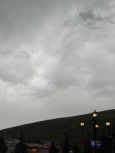 This image captures a cloudy day in Hrazdan, Armenia. The sky is filled with heavy, gray clouds, creating an overcast atmosphere. In the foreground, a dark green, tree-covered hill rises steeply. At the base of the hill, a few buildings with tiled roofs are visible, along with dark green coniferous trees. A decorative lamppost with four illuminated lamps stands to the right of the frame, casting a warm glow. Below the lamppost, a brightly lit sign with colorful letters spells out "TECNIC." The overall scene depicts a somewhat muted urban landscape against a dramatic, cloudy sky.