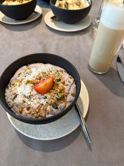 The image shows a table setting with a focus on a breakfast bowl. The bowl, filled with a creamy base and topped with granola, shredded coconut, seeds, passion fruit pulp, and a strawberry slice, sits on a white plate with a silver spoon. Adjacent to the bowl is a glass of layered milk or coffee drink. In the background, partially visible, are two additional bowls with similar contents, suggesting a group setting. The table surface appears to be covered in a grey tablecloth. The scene likely takes place in a cafe or restaurant in Нареченски бани, Bulgaria, though no external views confirm the specific location.