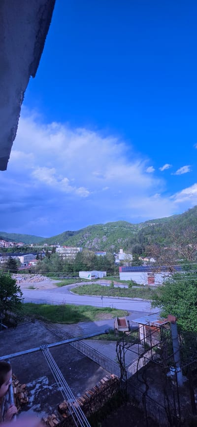 The image presents a view in Rudozem, Bulgaria. A mountain or steep hill, covered in green vegetation, dominates the background. The road winds along the base of the hill, and a few buildings and homes are visible. The sky is mostly blue with some clouds. In the foreground, there are parts of buildings and metallic structures from where the shot was taken.