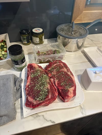 The image depicts a kitchen countertop in Saint-Jean-d'Aulps, France, prepared for cooking. Centrally positioned on a white rectangular platter are two raw, un-cooked ribeye steaks, each visibly seasoned with dried green herbs. To the left of the steaks, a tall glass jar of "Herbes de Provence" with a green label is present. Further left, a white bowl contains a chopped salad, primarily consisting of diced cucumber and red bell pepper. A folded grey dish towel lies partially visible on the far left.
Behind the steaks, a clear plastic container, covered with cling film, holds marinated meat pieces. Two additional glass jars are visible in the background: one with green olives, bearing a white and blue label indicating "ORIGINE FRANCE," and another with green contents, possibly pesto. To the right of the scene, a silver saucepan with a glass lid and grey silicone rim is situated. Adjacent to the saucepan, a white ceramic butter dish with its lid removed is visible, alongside an empty white rectangular serving dish. The countertop features a white surface with grey marble-like veining. The background consists of a dark grey or black splashback, with a section of a wooden cabinet frame visible on the right side. No individuals are present in the frame.