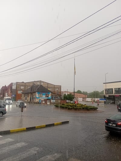 The image depicts a street scene in Tiachiv, Ukraine, during a heavy downpour. The road is flooded, with water pooling around vehicles and on the pedestrian crossing. Several cars are visible, navigating the wet conditions.
In the background, buildings line the street, some of which appear to be commercial establishments. One building has a sign that reads "ВІД 6 ВІКОН ПО ОПТОВІЙ ЦІНІ" (From 6 windows at wholesale prices). Another sign indicates "EVEREST-LAND LAND & REAL ESTATE". Further down the street, a sign with a blue arrow points to "УЖГОРОД ВОКЗАЛИ" (Uzhhorod Train Station). There are also signs for "CIAO!" and "FIMKA".
A prominent feature in the scene is a roundabout in the middle of the street, adorned with green bushes. A flagpole with a Ukrainian flag stands tall near the roundabout. The sky is overcast, indicating the continuing rain. The overall atmosphere is one of a typical rainy day in a Ukrainian town. No people are clearly visible due to the rain and distance. The time of day appears to be daytime, though the heavy clouds obscure direct sunlight.
