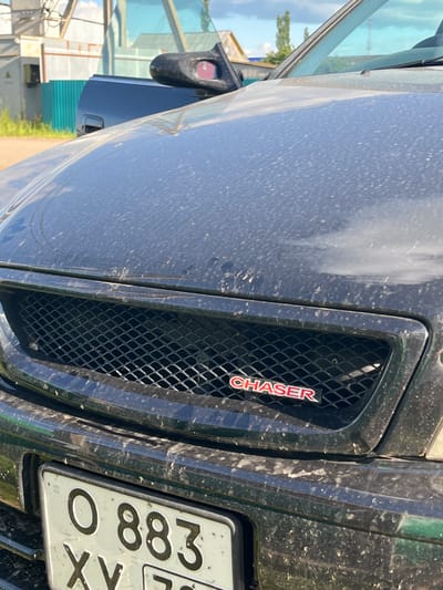 The image shows the front grille and hood of a black car, possibly a Toyota Chaser, covered in dust and small insects. A license plate with Cyrillic characters, "O 883 XY 72," is visible below the grille. In the background, there is a green fence, a building, and a utility pole. The scene appears to be outdoors on a sunny day. The car is parked on what looks like a dirt road or unpaved area.