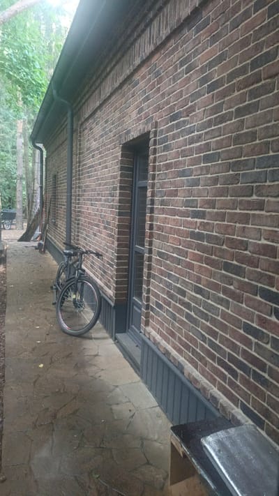 The image shows a section of a brick building with a gray door and a window. A bicycle is parked on a paved walkway in front of the building. The walkway is made of irregular flagstones. Trees and foliage are visible in the background, suggesting an outdoor setting. The overall scene appears to be a residential or possibly a commercial property in a semi-rural or suburban environment, consistent with locations like Nikolino, Russia. The lighting suggests it may be overcast or late in the day.