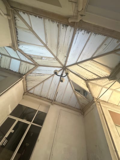 View looking up at an interior courtyard ceiling structure. The ceiling is constructed with a geometric pattern of metal framing and translucent or opaque panels, possibly polycarbonate or frosted glass, that have a ribbed texture. The panels are worn and appear dirty, with accumulated grime along the edges. A central light fixture with three frosted glass globes is suspended from the center of the ceiling. The courtyard walls are painted white or off-white, and a set of windows with a grid-like pattern is visible on one side. A pipe runs along the edge of the ceiling. The image may be from Santa Maria da Feira, Portugal.