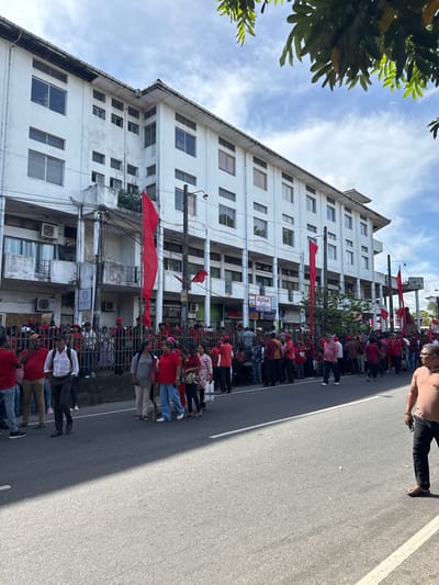 The image captures a street scene in Delkanda, Sri Lanka, showing a large gathering of people, predominantly dressed in red attire, lining a paved road. The perspective is tilted, making the multi-story white building on the left appear at an angle.
In the foreground and extending into the midground, a substantial crowd of individuals is visible, many wearing red shirts, indicating a possible organized event such as a rally or procession. Some red flags are held by participants, while others are affixed to utility poles along the street, reinforcing the impression of a collective gathering. One person in the immediate foreground, on the right side, wears a light brown shirt and dark trousers, holding a mobile phone, and appears to be observing or moving along the street.
The background features a multi-story, white-painted building with numerous rectangular windows. Utility poles with entangled wires run alongside the building. Green foliage from trees is visible in the upper left corner and intermittently along the street. Shadows cast on the road suggest it is daytime, under a clear sky with scattered wispy clouds.
Visible text on storefront signs along the building includes "Highland" followed by what appears to be "Bakery & Sweets". Another sign higher up on the building contains the word "SENANAYAKE" and possibly "Gems". The street itself is asphalt with white painted lines.