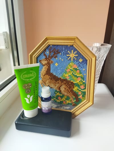 A framed cross-stitch artwork depicting a deer in a forest with a starry sky is displayed on a white surface next to a green tube of massage cream and a small dark blue bottle of essential oil. The framed artwork is octagonal with a wooden frame. The cross-stitch shows a brown deer standing in front of a green tree adorned with pink flowers, against a blue starry night sky. To the left of the artwork is a bright green tube with the brand name "ERSAG" in white lettering and a green leaf logo. The tube is labeled "CLEANBALL MASAJ KREMİ". Next to the cream tube is a small, dark blue glass bottle with a white cap, also with "ERSAG" branding, labeled "Sırt ve Boyun" and "Lavanta Yağı". Both the cream tube and the essential oil bottle are placed on a black rectangular object that appears to be a portable power bank. In the background, to the right of the framed artwork, is a clear glass vase with a textured surface. The scene is lit by natural light coming from the left, suggesting it might be near a window. The wall behind the objects has a peachy-pink hue. The overall setting appears to be an indoor display on a windowsill or a table near a window. There is no indication of the location being in Dolyna, Ukraine, or any other specific city.
