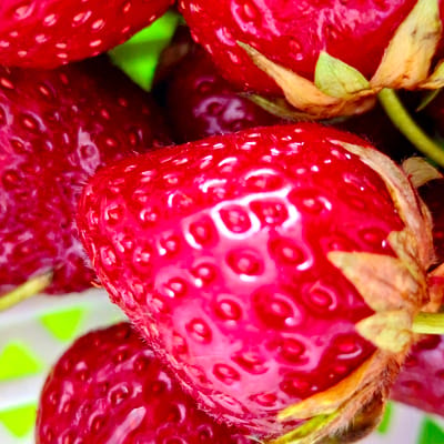 The media file is a close-up, macro photograph of several ripe strawberries. The strawberries are a vibrant red and their textured surfaces, dotted with seeds, are clearly visible. Some of the strawberries are partially obscured by others, creating a sense of depth and abundance. The focus is tight on the fruit, with the background blurred and showing hints of green and white, possibly indicating packaging or foliage. The lighting highlights the glossy sheen of the strawberries, suggesting they are fresh and possibly wet, perhaps from washing or dew. The image captures the intricate details of the strawberries' skin and the small green stems and calyxes where they were attached. There is no discernible activity or event taking place; the image is a still life composition focusing solely on the appearance of the fruit. No text is visible in the image. The location context of Baden-Baden, Germany does not provide any specific visual cues within the image itself.