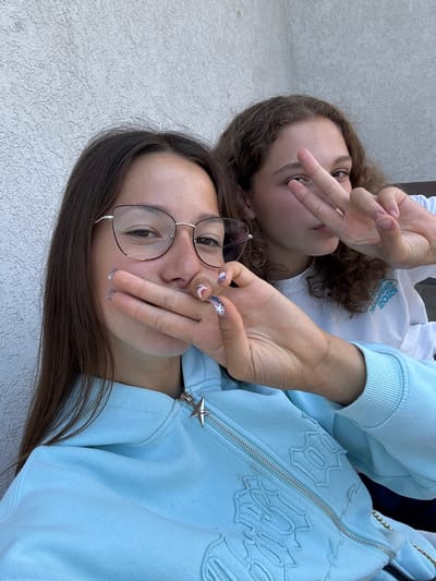 Two young women are posing for a selfie against a textured light-colored wall. The woman in the foreground is wearing glasses and a light blue zip-up hoodie with a star zipper pull. Her right hand is covering her mouth, and her left hand, adorned with decorative nails, rests on her chest. The woman in the background has curly brown hair and is wearing a white sweatshirt. She is making a peace sign with her right hand in front of her face. The image appears to be taken outdoors, possibly on a balcony or patio, given the wall texture and natural lighting. No specific landmarks identifying Bozhurishte, Bulgaria, are visible in the image.