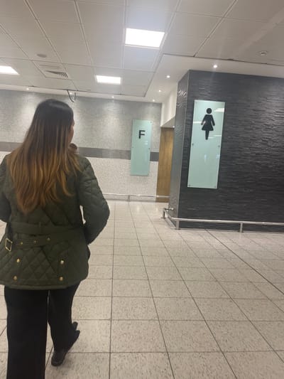 An indoor, vertically oriented image captures a person from behind, standing in what appears to be a public building, possibly a travel hub or shopping center in Horley, United Kingdom. The person has long, brown hair with lighter highlights, wearing a dark green quilted jacket with a belted waist and dark trousers or leggings paired with dark boots. They are facing away from the camera, looking towards a dark, textured wall.
On this dark wall, two rectangular, frosted glass signs are mounted. The upper sign displays a black female pictogram, typically indicating a women's restroom. Below it, a second, similar sign features a large capital letter "F". Both signs likely have unreadable smaller text or braille next to their main symbols.
The floor is covered in light-colored, patterned tiles, while the ceiling consists of square white panels with embedded rectangular and circular light fixtures, providing bright artificial illumination. A metal railing is visible on the right side of the frame. The overall setting suggests a modern, well-lit, and functional public space, with the signs clearly directing individuals, likely to a specific floor or facility such as a female restroom.
