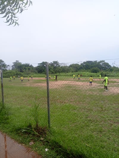 The image depicts a group of young people playing soccer on a dirt field, likely in Ife, Nigeria. The game appears to be a casual match, with several players scattered across the field, some actively engaged with the ball while others are observing or positioning themselves. The players are mostly dressed in bright yellow or lime green soccer jerseys and shorts, suggesting they are part of a team or a training session. One player in a lime green jersey stands out as he is facing the camera, positioned in the foreground of the field.

The scene is set in an outdoor environment with a chain-link fence in the foreground, partially obscuring the view of the field. Beyond the field, a dense line of lush green trees and vegetation forms the backdrop, characteristic of a tropical or sub-tropical climate. The sky is overcast with a light, hazy quality, indicating a cloudy day, possibly with the threat of rain. The ground on the field is predominantly reddish-brown dirt, with patches of sparse green grass around the edges. There are puddles of water on the ground near the fence, suggesting recent rainfall. The overall atmosphere is one of everyday activity in a community setting. There is no visible text in the image.
