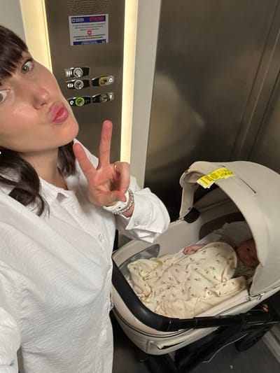 A woman is taking a selfie with a baby in a stroller inside an elevator. The woman, wearing a white shirt, is making a peace sign with her left hand. The baby is wrapped in a blanket with a floral pattern inside a white stroller. The elevator has a metal panel with buttons for different floors and informational signs. The location is unknown, and there is no indication of Cinisi, Italy.