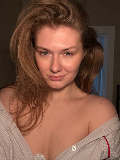 A close-up portrait depicts an adult female positioned indoors in Tashevka, Russia. The subject faces forward with a slight smile and direct gaze. She has fair skin, light blue-green eyes, natural eyebrows, and long, wavy reddish-brown hair that frames her face and shoulders. Her hair appears slightly voluminous. She is wearing a light-colored, possibly grey or white, striped shirt with red piping visible on the right side, worn unbuttoned and off both shoulders, exposing her upper chest and clavicle area. The background is dimly lit and out of focus, with a faint vertical light-colored doorframe or trim visible on the right side. The overall lighting is soft, illuminating the subject from the front.