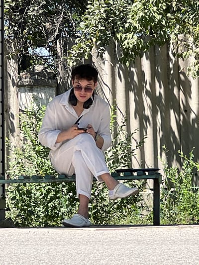 A young man in white clothing and sunglasses sits on a green metal bench in an outdoor setting. He is wearing black headphones around his neck and holding a mobile phone in his hands, seemingly engaged with it. The bench is situated in front of a lush green hedge, and behind the hedge is a grey corrugated fence with shadows cast upon it from overhead foliage. The background also includes some stone structures and dense green leaves from trees. The lighting suggests it is daytime. The location is identified as Дачное, Kyrgyzstan.