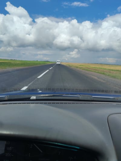 An eye-level, point-of-view shot from inside a vehicle driving on a paved road. The road has white lane markings and extends into the distance. On either side of the road are fields of green and yellow grass. The sky is blue with large, fluffy white clouds. A white van is visible in the distance, driving in the same direction as the camera. The dashboard of the vehicle is visible in the foreground, with a speedometer showing a speed of approximately 160 km/h. The scene is likely in rural Belarus, possibly within the Azdzelinski Sel'ski Savet region, given the flat, open landscape and type of road.