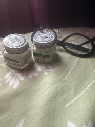 The image displays two small white jars with metal lids, likely containing a balm or ointment, placed on a light green tablecloth with a subtle floral pattern. One jar is positioned in the foreground to the left, and the other is slightly behind and to the right. The lids of both jars feature intricate circular designs with green accents and text in an Asian script. Behind the jars, a pair of black-framed eyeglasses lies on the tablecloth, with their lenses partially obscuring the second jar. In the background, a dark, out-of-focus surface, possibly fabric, is visible. The lighting suggests an indoor setting. There is no discernible information about the location in the city of Tatarasaya Pishly, Russia.