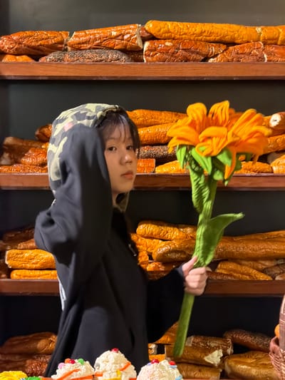 A person with a tan and black camouflage-patterned hat and a black jacket is holding a decorative orange flower against a background of wall-mounted bread-like sculptures. The location is Thuận Hóa Ward, Vietnam.