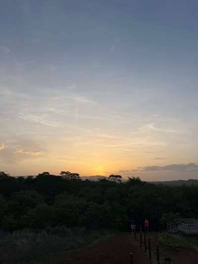 This is a landscape photograph taken at sunset in Nindirí, Nicaragua. The sky transitions from a pale blue at the top to hues of orange and yellow near the horizon, with scattered clouds. In the foreground, a dark path made of earth leads downwards, lined with red posts. Two individuals, one wearing a blue shirt and white shorts, and another in an orange shirt and dark pants, are walking away from the camera down the path. To the right of the path, there is a white railing and fence. Behind the path and people, a dense forest of dark green trees stretches across the landscape, with rolling hills visible in the distance. The overall impression is one of a tranquil natural setting at dusk.