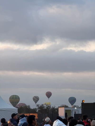 The image depicts several hot air balloons floating in the sky above San Francisco Mazapa, Mexico. People are gathered on the ground, looking up at the balloons. Some are taking pictures. The sky is cloudy. There's a stage and some temporary structures nearby, suggesting it's an event.