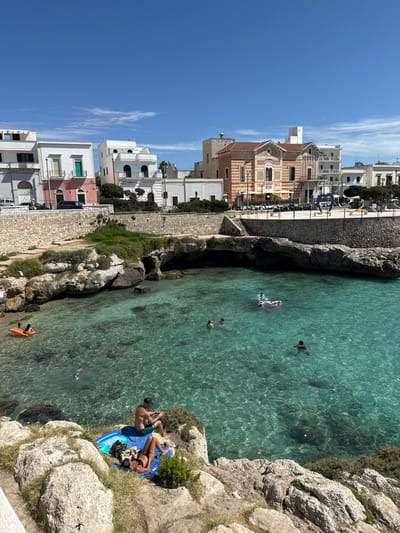 The image depicts a sunny coastal scene at Santa Caterina di Nardò, Italy, specifically a natural rock-formed cove. The sky is clear blue with minimal cirrus clouds. The water in the cove is transparent and turquoise, revealing the seabed. The cove is surrounded by light-colored rocky cliffs and outcroppings, with sparse green vegetation. Several small natural caves or overhangs are visible along the waterline.

In the foreground, on the rocks, two individuals are present. A male, wearing dark shorts and a dark cap, is seated on a blue and white striped towel, facing the water, appearing to interact with a small object in his hands. Adjacent to him, a female wearing a dark bikini is lying prone on a blue towel, sunbathing.

In the water, approximately five to six individuals are visible. One person is on an orange inflatable raft near the left edge, while another is on a pink and white inflatable further back on the right. Other individuals are swimming or wading in the clear water, with one person partially submerged in the lower left.

In the background, a town skyline extends above a stone retaining wall. Buildings are predominantly white or light-colored, with a multi-story building featuring a distinct ornate facade of terracotta and striped patterns dominating the center-right. Other buildings include one with a pink-toned lower section and a teal door. A paved area or plaza is visible behind the retaining wall, containing parked cars and scooters, as well as a large dark sculpture or structure. Stone steps lead down from the town level towards the water on the right.