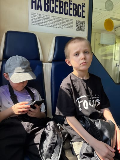 Inside a public transport vehicle, likely a train, in Vyatskiye Polyany, Russia, two young boys are seated. The boy on the left wears a grey baseball cap, a light purple and white long-sleeved shirt with an "E" logo, and dark pants. He is looking down intently at a black smartphone he holds with both hands. A black strap from a bag or device crosses his chest. To his right, the second boy wears a black t-shirt with "MODEL MANAGEMENT Focus" printed in white, and light-colored shorts. He looks directly at the viewer with a neutral expression. A black, textured, possibly faux leather bag rests on his lap. Strong sunlight enters from the right, casting distinct shadows across the interior. The seats are blue with white trim. Above the boys, a white poster in Cyrillic advertises "НА ВСЕВ СВЁ" and details a "SUPER SMART PROMO" credit offer from "POCHTA BANK," including a QR code. Through the window on the right, blurred green foliage is visible, indicating movement or a natural setting. A yellow, circular object is adhered to the window.