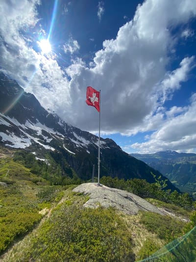 A vibrant outdoor shot captures a mountainous alpine landscape under a partly cloudy sky, with a bright sun in the upper left quadrant emitting prominent rays and lens flare. The scene is located near Innertkirchen, Switzerland. A red Swiss flag, featuring a white cross, is prominently displayed on a metallic flagpole positioned on a large, flat, light-colored rocky outcrop in the midground, slightly to the right of the frame's center. The flag is unfurled towards the left. The foreground consists of lush green alpine vegetation, including grasses and various shrubs, traversed by a narrow dirt path visible on the left. To the left, a large mountain range rises steeply, showcasing patches and streaks of snow against dark rock faces and lower green slopes. In the far background, additional green mountainous terrain and a valley are visible. The sky is a mix of deep blue and scattered white and grey cumulus clouds. No human subjects are present in the image.