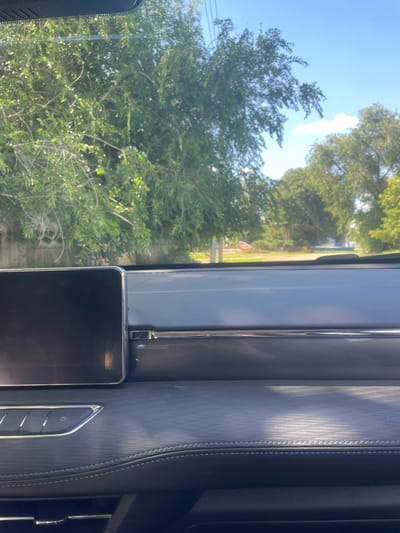The image shows a view from inside a car dashboard looking out through the windshield. The dashboard is made of dark material with textured accents and a large screen on the left. Outside, there are trees with green leaves and a blue sky with a few clouds. A road is visible in the distance, with some buildings and greenery on either side. The scene appears to be in a suburban or rural area. The location of Линёвка, Russia is not discernible from the image.